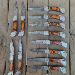 May include: An assortment of folding knives with ornate silver-colored bolsters and handles featuring a combination of orange and brown wood. The blades have a textured pattern. The knives are arranged on a wooden surface.