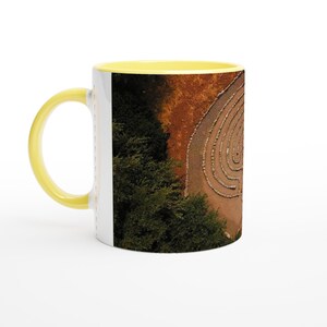 May include: A yellow and white ceramic mug featuring a photograph of a stone labyrinth set within a forest. The labyrinth is surrounded by trees and brown earth.