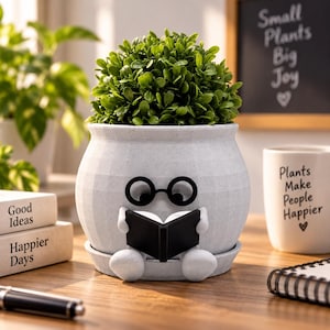 May include: A light gray, textured, round planter featuring a character with black glasses reading a black book. The planter holds a vibrant green, bushy plant and sits on a matching saucer. The design includes small white arms and feet.