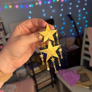 Peut inclure: Une boucle d'oreille pendante dorée composée de deux grandes étoiles superposées verticalement. Les étoiles sont ornées de petites breloques en forme d'étoile et de minuscules cristaux ronds bleus, violets et transparents suspendus à de fines chaînes.
