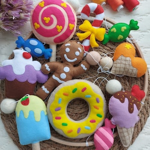 Puede incluir: Una colección de adornos de comida de fieltro coloridos, que incluyen una piruleta rosa, un hombre de jengibre, una rosquilla y un cono de helado. Otros adornos incluyen un bastón de caramelo, un caramelo azul y una magdalena. Los adornos están dispuestos en una bandeja tejida.