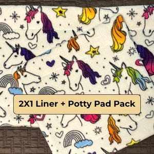 May include: A white fabric liner and potty pad set featuring a repeating pattern of colorful unicorns, rainbows, hearts, and stars. The text 2X1 Liner + Potty Pad Pack is printed on a tan rectangular label centered on the fabric surface.