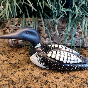 May include: A hand-painted wooden loon figurine. The bird has a black head and neck, a long black beak, and a black body with white spots. The loon has red eyes and is set against a blurred background.