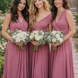 May include: Three women in matching mauve-colored bridesmaid dresses, each holding a bouquet of white roses and greenery. The dresses have a V-neck design. The women are smiling, posing outdoors in a garden setting.