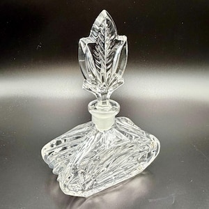 May include: Clear glass perfume bottle with a leaf-shaped stopper. The bottle has a square base with a textured, ribbed design. The stopper is a detailed leaf shape, adding a decorative element to the vintage fragrance container.