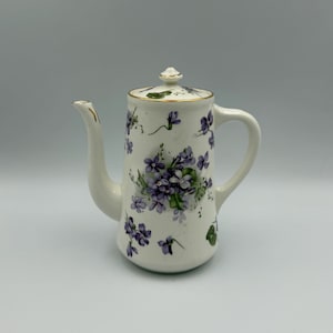 May include: A white ceramic coffee pot with a lid and handle, decorated with purple floral patterns and gold trim. The pot features a slender spout and a rounded body, perfect for serving coffee or tea.