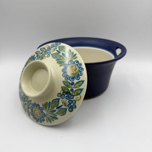 May include: A deep blue ceramic bowl with a matching lid. The lid features a cream-colored background decorated with a repeating pattern of blue and green floral and leaf motifs. The bowl has two integrated side handles and a smooth finish.