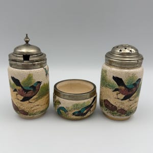 May include: A set of three ceramic kitchen items: a lidded jar, a small open bowl, and a shaker. Each piece is decorated with a painted bird design in shades of blue, brown, and green, with a silver-colored metal lid.