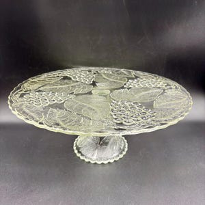 May include: A clear glass cake stand with a decorative grape and leaf design. The stand has a round, scalloped edge and a pedestal base. The design is etched into the glass, creating a textured surface. The stand is suitable for displaying cakes or other desserts.