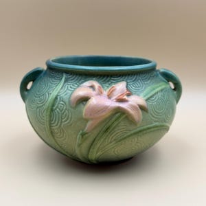May include: A round ceramic bowl with two small handles, featuring a matte green finish and a raised, embossed light pink lily flower design. The surface is decorated with a repeating wave-like pattern that covers the exterior of the vessel.