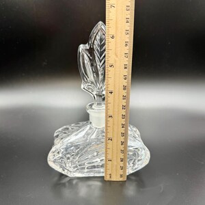 K&ouml;nnte beinhalten: Eine klare Glasparf&uuml;mflasche mit einem blattf&ouml;rmigen Verschluss. Die Flasche ist etwa 13 cm hoch und hat ein dekoratives, strukturiertes Design. Der Verschluss hat die Form eines Blattes. Die Flasche steht auf dunklem Hintergrund.