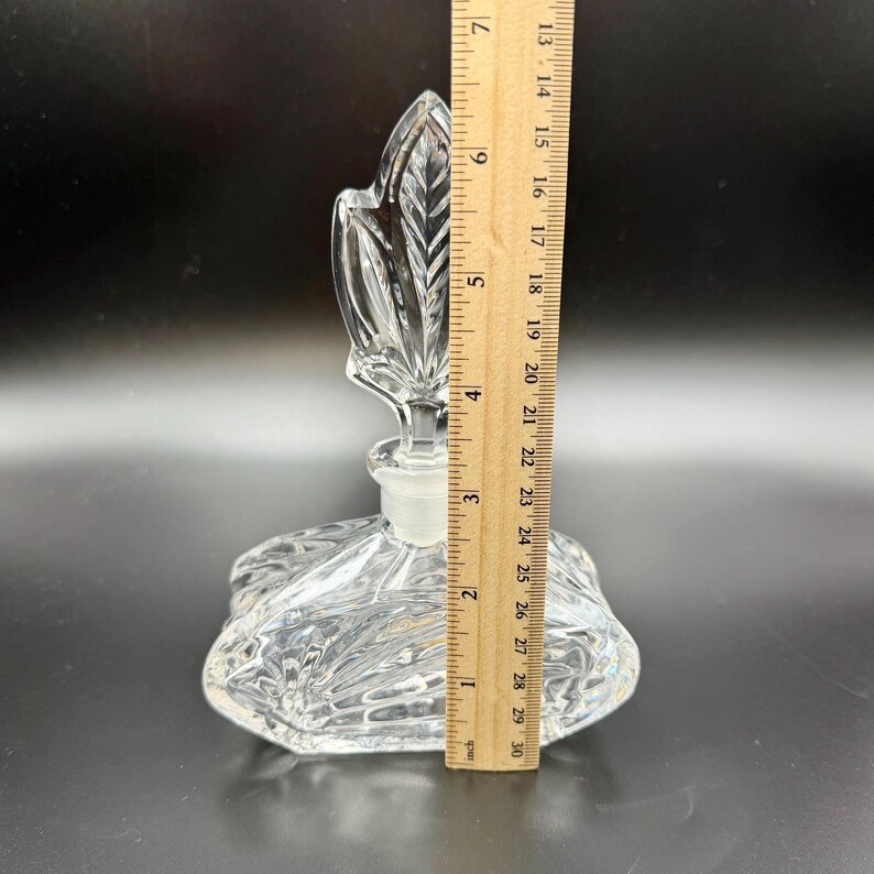 K&ouml;nnte beinhalten: Eine klare Glasparf&uuml;mflasche mit einem blattf&ouml;rmigen Verschluss. Die Flasche ist etwa 13 cm hoch und hat ein dekoratives, strukturiertes Design. Der Verschluss hat die Form eines Blattes. Die Flasche steht auf dunklem Hintergrund.