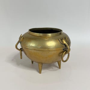 May include: A round brass vessel with a wide opening, featuring three pointed legs and two side handles shaped like stylized animal heads with attached rings. The metal surface has a polished gold-toned finish with visible texture.