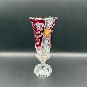 May include: A clear crystal vase with a ruby red interior and intricate floral and geometric cut patterns. The vase has a flared top, a pedestal base, and a gold-colored band near the base. A small sticker is affixed to the side.
