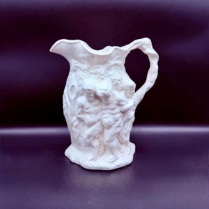 May include: A white ceramic pitcher with a detailed relief design. The pitcher features a sculpted handle and a decorative relief of figures, grapes, and foliage. The pitcher has a ruffled rim and a wide base. The overall design is classical.