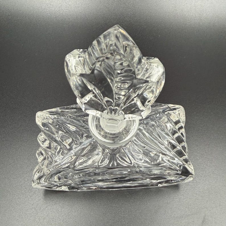 K&ouml;nnte beinhalten: Eine klare Parf&uuml;mflasche aus Glas mit einem blattf&ouml;rmigen Verschluss. Die Flasche hat einen quadratischen Sockel mit einem strukturierten, geometrischen Design. Der Verschluss ist ebenfalls klar und hat ein detailliertes, stilisiertes Blattdesign.