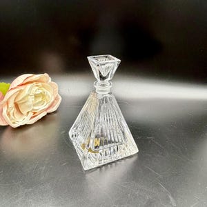 May include: A clear, geometric-shaped glass decanter with a square stopper. The decanter has vertical lines etched into the glass, creating a textured appearance. A pink and cream-colored rose is in the background.
