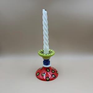 May include: A white spiral taper candle sits in a ceramic candlestick holder. The holder features a lime green top rim, a blue middle section with yellow zigzag lines, and a wide red base decorated with black-outlined green and yellow spots.