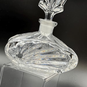 K&ouml;nnte beinhalten: Klarer Parf&uuml;mflakon aus Glas mit dekorativem Verschluss. Die Flasche hat einen abgerundeten Boden und ein strukturiertes, f&auml;cherartiges Muster. Der Verschluss ist ein klares, verziertes Design. Die Flasche wird auf einem transparenten St&auml;nder ausgestellt.