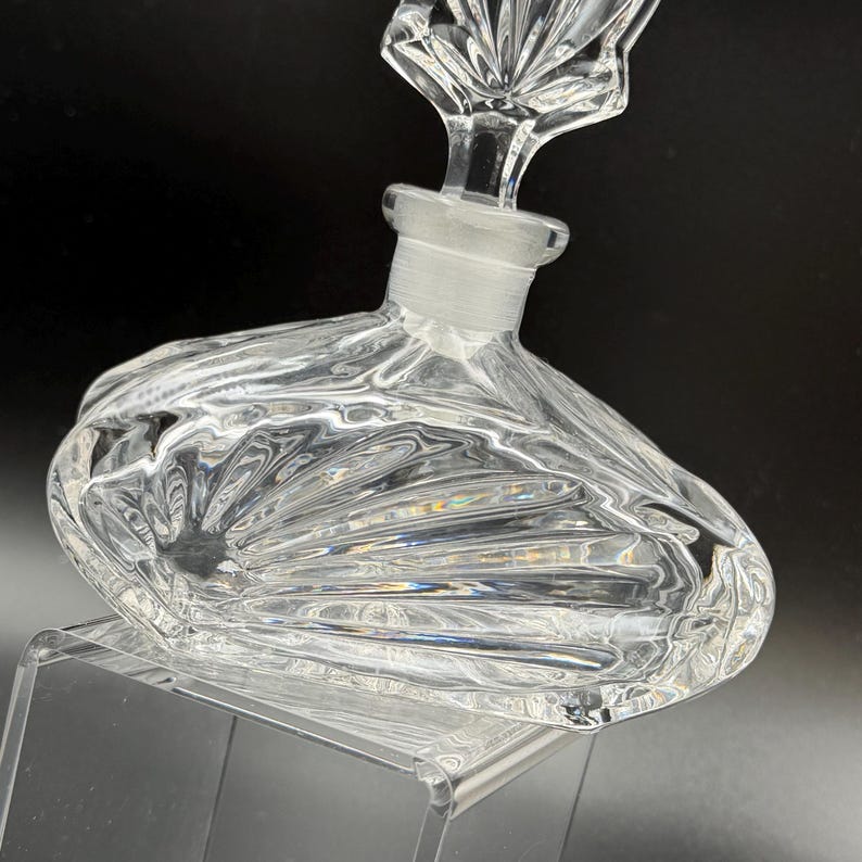 K&ouml;nnte beinhalten: Klarer Parf&uuml;mflakon aus Glas mit dekorativem Verschluss. Die Flasche hat einen abgerundeten Boden und ein strukturiertes, f&auml;cherartiges Muster. Der Verschluss ist ein klares, verziertes Design. Die Flasche wird auf einem transparenten St&auml;nder ausgestellt.