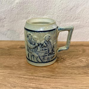 May include: A stoneware beer mug with a detailed relief scene of figures around a barrel. The mug has a light blue-gray color with darker blue accents and a sturdy handle. The design is reminiscent of traditional German beer steins.