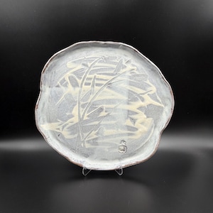 May include: A handcrafted, irregularly shaped ceramic plate with a light gray glaze. The plate features a cream-colored botanical design and a small, stamped emblem. The rim has a natural, unglazed brown edge.