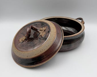 May include: A handmade, open ceramic bowl with a lid. The bowl is dark brown with a lighter brown rim and a lid with a handle and decorative leaf and grape design. The bowl is on a white background.