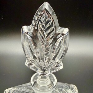 K&ouml;nnte beinhalten: Klarer Glasflaschenverschluss mit Blattform. Der Verschluss hat oben eine detaillierte, strukturierte Blattform mit abgerundeter Basis. Das Glas ist transparent, l&auml;sst Licht durch und ist vor einem dunklen Hintergrund platziert.