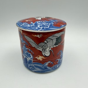 May include: A cylindrical ceramic lidded container featuring a vibrant red background decorated with blue and white stylized ocean waves and a large white crane with black wing details and a red crown flying across the side and lid surface.