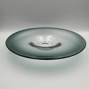 May include: A large, shallow, teal-colored glass bowl with a wide, flat rim and a central depression. The bowl has a smooth, reflective surface and a dark rim. The design is simple and modern, suitable for decorative or functional use.