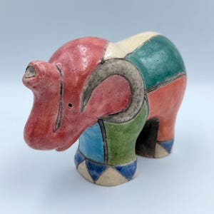 May include: A ceramic elephant figurine featuring a segmented design with a color palette of red, green, blue, and cream. The surface is divided by dark outlines, creating distinct geometric shapes across the body, head, and ears of the animal.