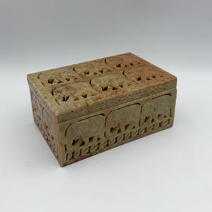 May include: A rectangular, beige stone box with intricate carvings of elephants. The lid and sides feature detailed elephant designs, creating a decorative storage piece. The box is made of a light-colored stone.