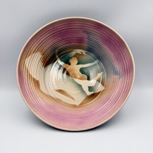 May include: A round ceramic bowl featuring a ribbed interior texture. The surface displays a gradient of purple, blue, and tan hues. A white silhouette of a figure in a dynamic, reaching pose is centered within the bowl, contrasting against the colors.
