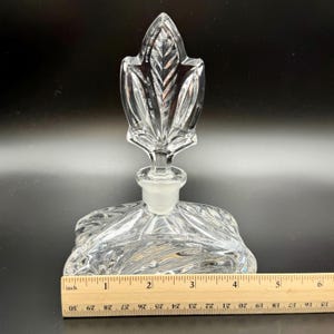 K&ouml;nnte beinhalten: Klarer Glasparf&uuml;mflakon mit blattf&ouml;rmigem Verschluss. Die Flasche hat einen quadratischen Boden mit strukturiertem Design. Der Verschluss ist etwa 10 cm hoch. Die Flasche steht auf schwarzem Hintergrund.
