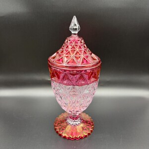 May include: A decorative glass jar with a lid, featuring a vibrant pink and clear diamond-cut pattern. The vessel has a pointed finial on top, a tapered body, and a scalloped circular base, all crafted from pressed glass with a geometric texture.