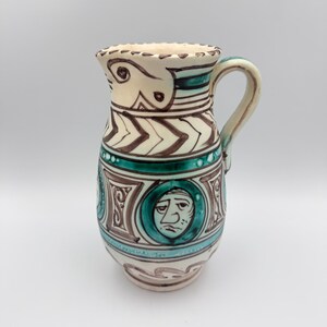 May include: A ceramic pitcher featuring a cream-colored base with hand-painted dark brown and teal green decorative patterns. The design includes a series of circular medallions containing stylized human faces, chevron bands, and a curved handle.
