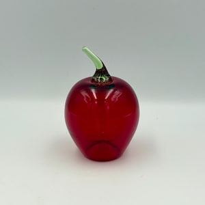 May include: A decorative glass apple with a vibrant red body and a green stem. The apple is smooth and glossy, reflecting light. This handcrafted piece is a unique home decor item.