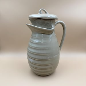 May include: A light gray ceramic pitcher with a glossy finish, featuring a ribbed, horizontal texture along the body, a curved handle, a wide pouring spout, and a matching lid with a small, looped handle on top for easy removal and handling.