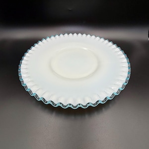 May include: A white glass plate with a ruffled edge and a light blue trim. The plate has a circular center and a scalloped edge. The plate is on a black surface.