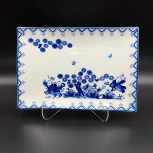 May include: A rectangular white ceramic serving tray featuring a blue decorative border and a central illustration of children playing among stylized flowers, foliage, and small butterflies. The design is rendered in a classic blue and white color palette.