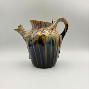 May include: A ceramic pitcher with a unique, organic design. The pitcher features a spout and handle, with a color palette of brown, blue, and gold. The surface has a textured, glazed finish, adding depth and visual interest to the piece.