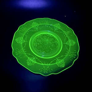 May include: A glowing green glass plate with a scalloped edge. The plate features embossed floral patterns around the rim and center. The plate is illuminated with a black light, causing it to fluoresce brightly against a dark background.