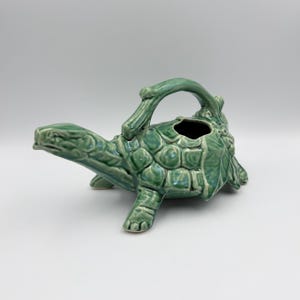 May include: A glazed ceramic teapot shaped like a turtle with a glossy sage green finish. The handle is designed to resemble a branch, while the turtle's shell features a textured, segmented pattern. The spout extends from the turtle's head.
