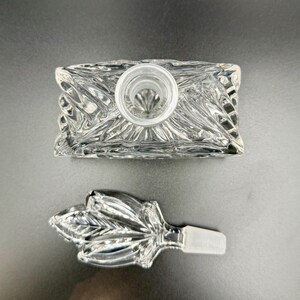 K&ouml;nnte beinhalten: Eine rechteckige, transparente Glasparf&uuml;mflasche mit einem dekorativen, blattf&ouml;rmigen Verschluss. Die Flasche hat ein strukturiertes, geschliffenes Glasdesign. Der Verschluss ist abgenommen und zeigt die &Ouml;ffnung der Flasche. Der Hintergrund ist einfarbig dunkelgrau.