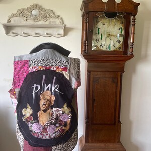 May include: A patchwork vest with a black graphic featuring the word "pink" and an image of a singer. The vest is displayed on a mannequin. A vintage grandfather clock and a decorative wall shelf are in the background.