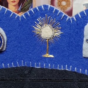 May include: A blue felt religious display with white stitching. It features a silver pendant of the Virgin Mary, a gold monstrance, and a blue and silver cross pendant. The background is dark.