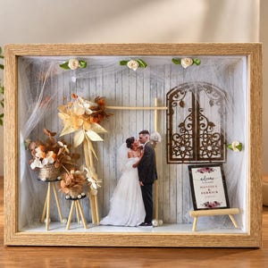 May include: A shadow box frame containing a miniature wedding scene with a cutout of a bride and groom, dried flowers, a wooden arch, a decorative metal window cutout, and a small sign that reads Welcome to the wedding of Monique & Derrick.