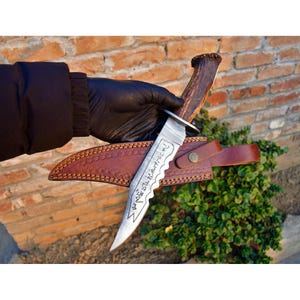 May include: A fixed-blade knife with a serrated spine and etched runic symbols on the silver-toned metal blade. The handle is crafted from textured brown antler, and the knife is accompanied by a brown leather sheath with decorative stitching.