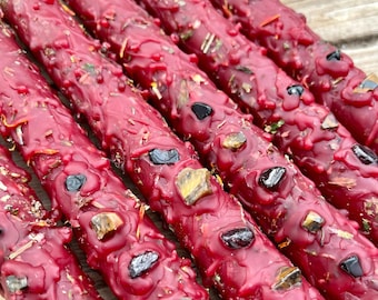 May include: A close-up of several red candle sticks with embedded crystals and herbs. The candles are arranged in a row, with the crystals and herbs visible on the surface of the candles.
