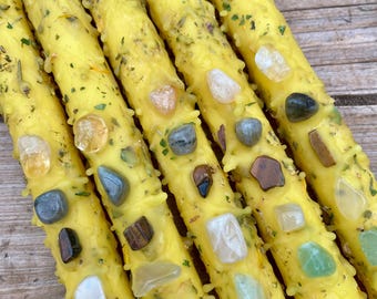 Puede incluir: Cinco velas amarillas con cristales y hierbas incrustados. Las velas están dispuestas en fila, con los cristales y las hierbas visibles en la superficie de las velas.